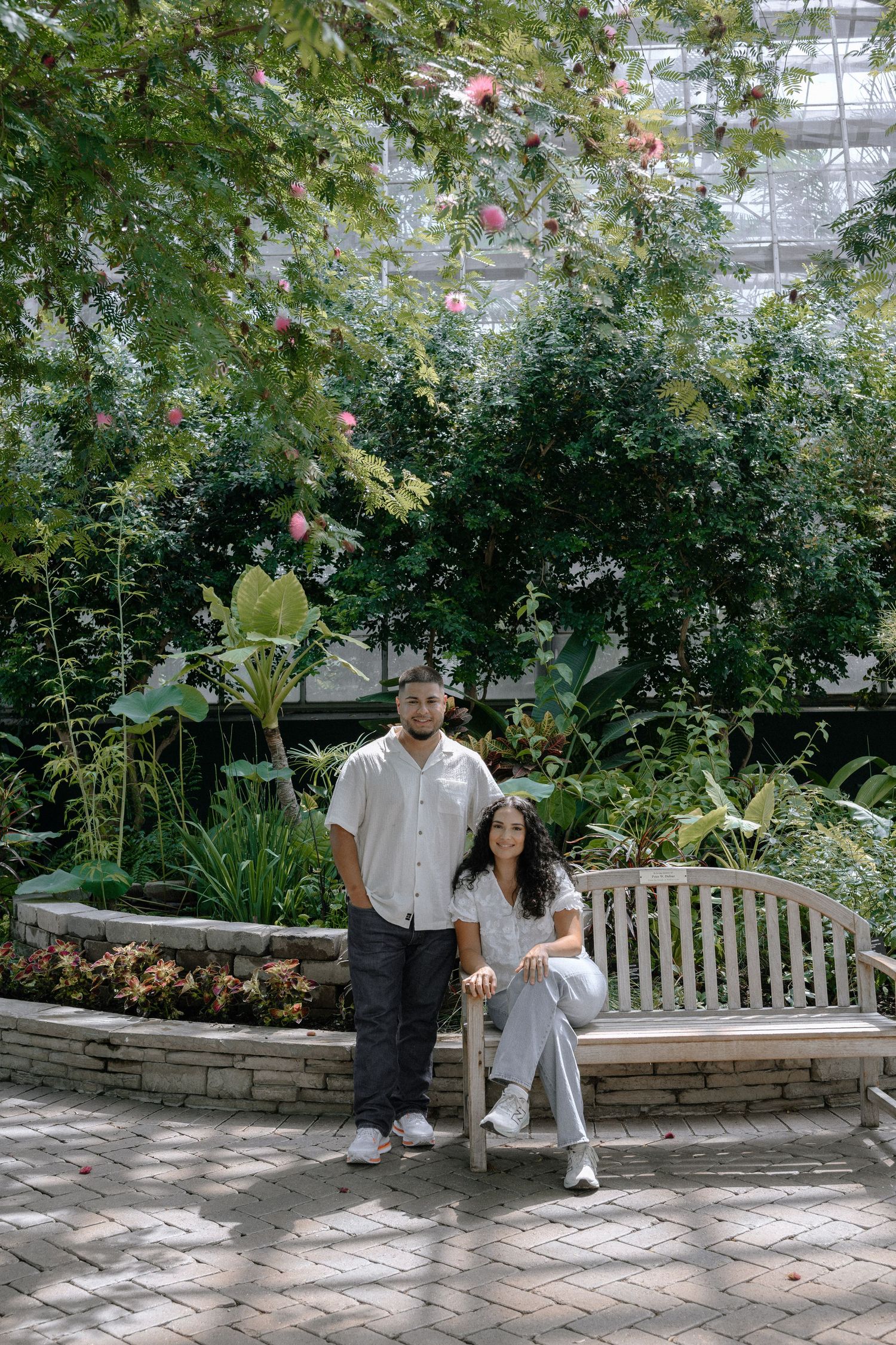 Two people standing together on a brick path inside a greenhouse with lush ferns and tropical plants surrounding them.