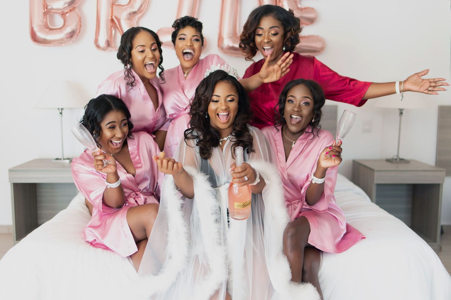 Bridesmaids in pink robes pose playfully with bride in white on bed at pre-wedding celebration party.
