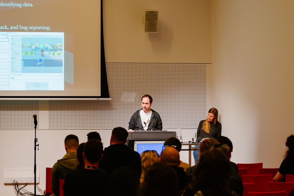 A presenter speaks at a podium while audience members listen in a lecture hall with a projected screen display.