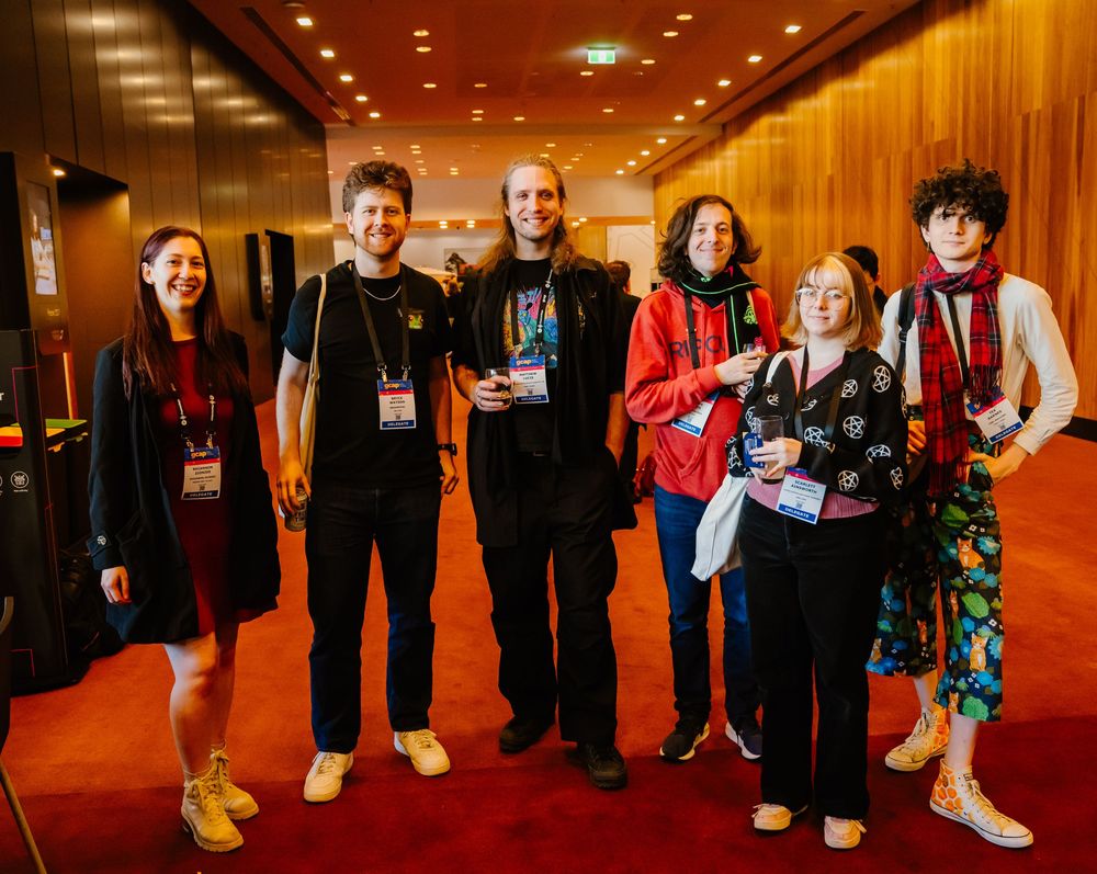 A group of six attendees wearing conference badges pose together in a warmly lit hallway with wood paneled walls.