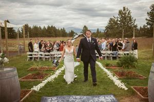 A couple walks down a flower-lined aisle at an outdoor wedding ceremony with guests seated on white chairs.