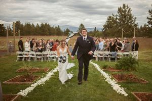 An outdoor wedding ceremony setup features white chairs and floral petal aisle decorations on a grassy field.