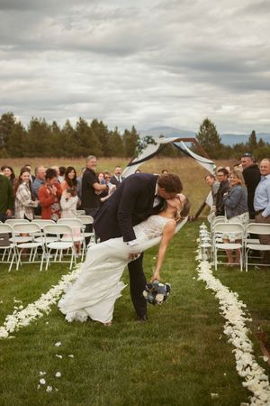 A wedding couple performs a dramatic dip at an outdoor ceremony with white chairs and floral petal aisle decorations.