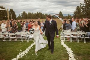A wedding ceremony concludes as a couple walks down a petal-lined aisle with guests in white chairs on a grassy lawn.