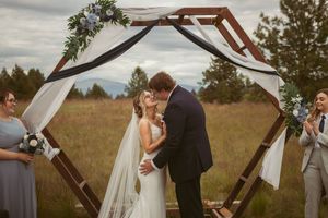 Rustic wooden arch decorated with greenery and white fabric stands in an outdoor wedding setting.