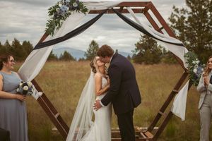 A romantic moment under a wooden hexagonal arch decorated with white fabric at an outdoor wedding.