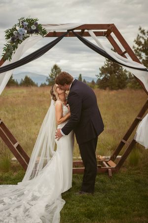 A romantic wedding moment captured under a wooden arch decorated with flowing white fabric.