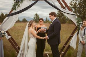 Wedding ceremony under a wooden hexagonal arch with flowing white fabric in a natural field setting.