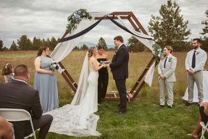 Outdoor wedding ceremony under rustic wooden arch with wedding party and guests gathered on grassy field.