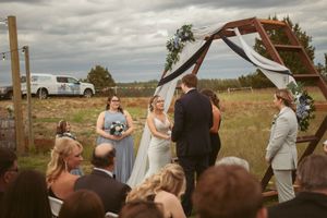 An outdoor wedding ceremony takes places under a rustic wooden arch with white draping.