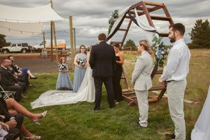 An outdoor wedding ceremony takes place under a wooden arch with guests seated on white chairs.