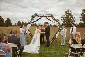 A ceremony unfolds beneath a rustic wooden arch decorated with white fabric on a sunny outdoor lawn.