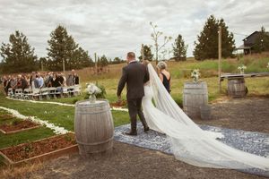A trailing wedding veil flows behind as couple walks past wine barrels at outdoor ceremony.