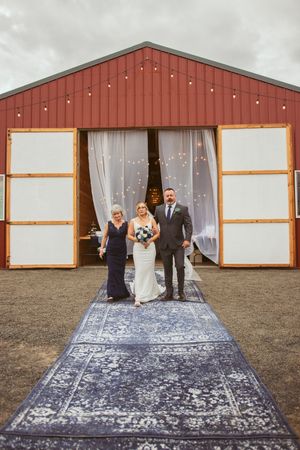 Wedding party stands at entrance of rustic red barn with ethereal white curtains and vintage blue rug leading to doorway.