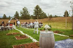 Outdoor country wedding ceremony setup with white flower petals, wine barrels and wooden chairs on grassy hillside.