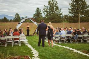Wedding guests seated in white chairs along a petal-lined aisle leading to a wooden ceremony arch.