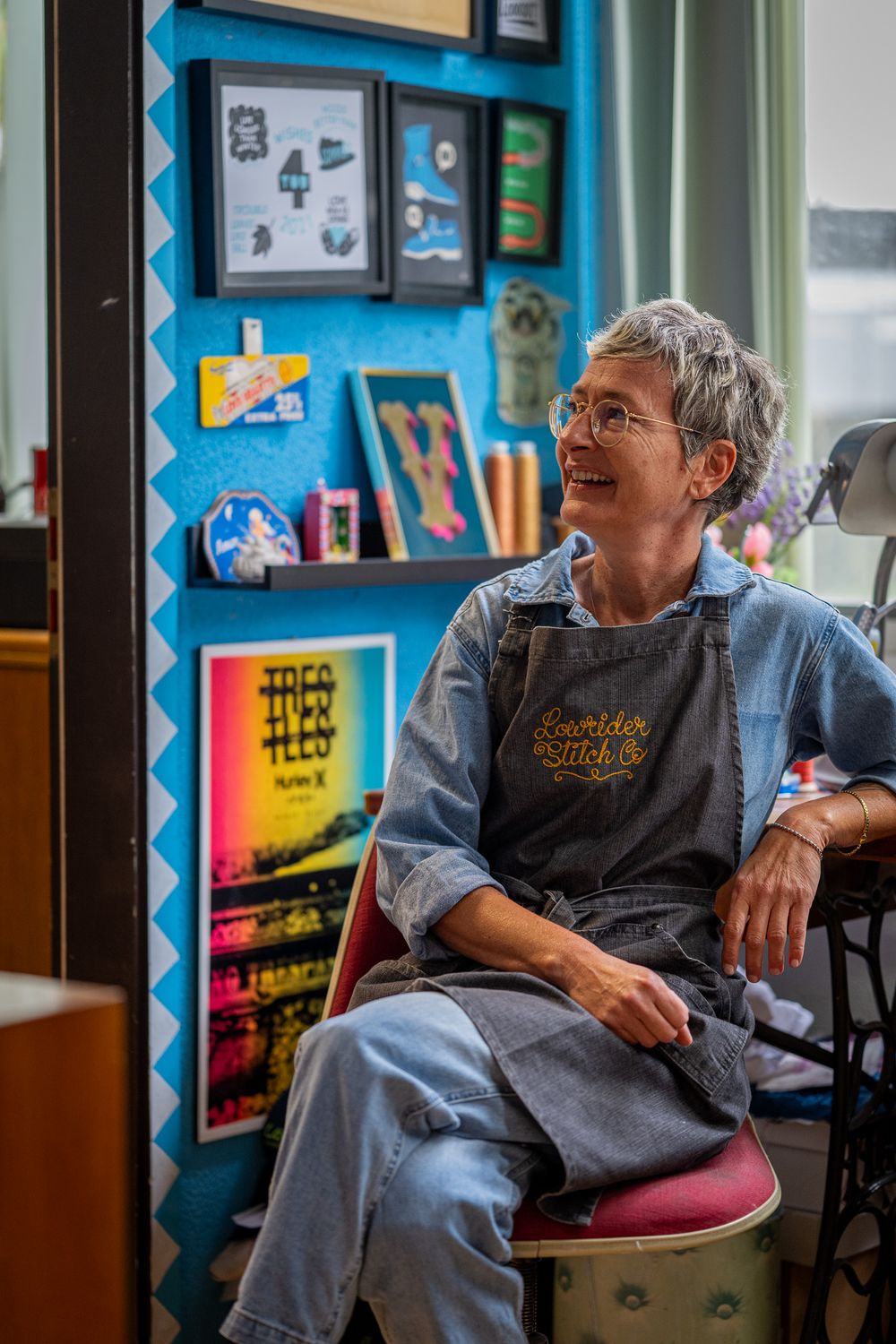 Someone in blue denim overalls sits casually in a room with colorful wall displays and art in the background.