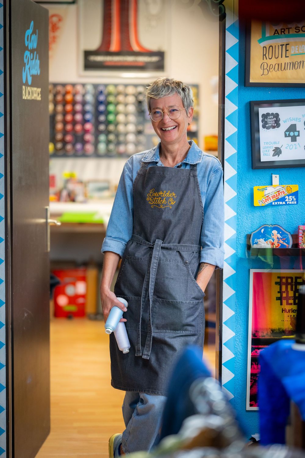 Staff member in blue shirt and gray apron smiles warmly while working in colorful retail store environment.