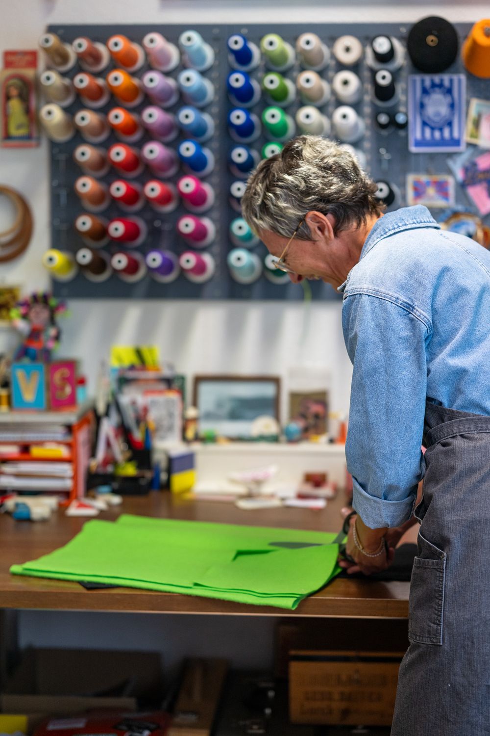 Person in denim apron working at a craft station with rows of colorful thread spools displayed on the wall.