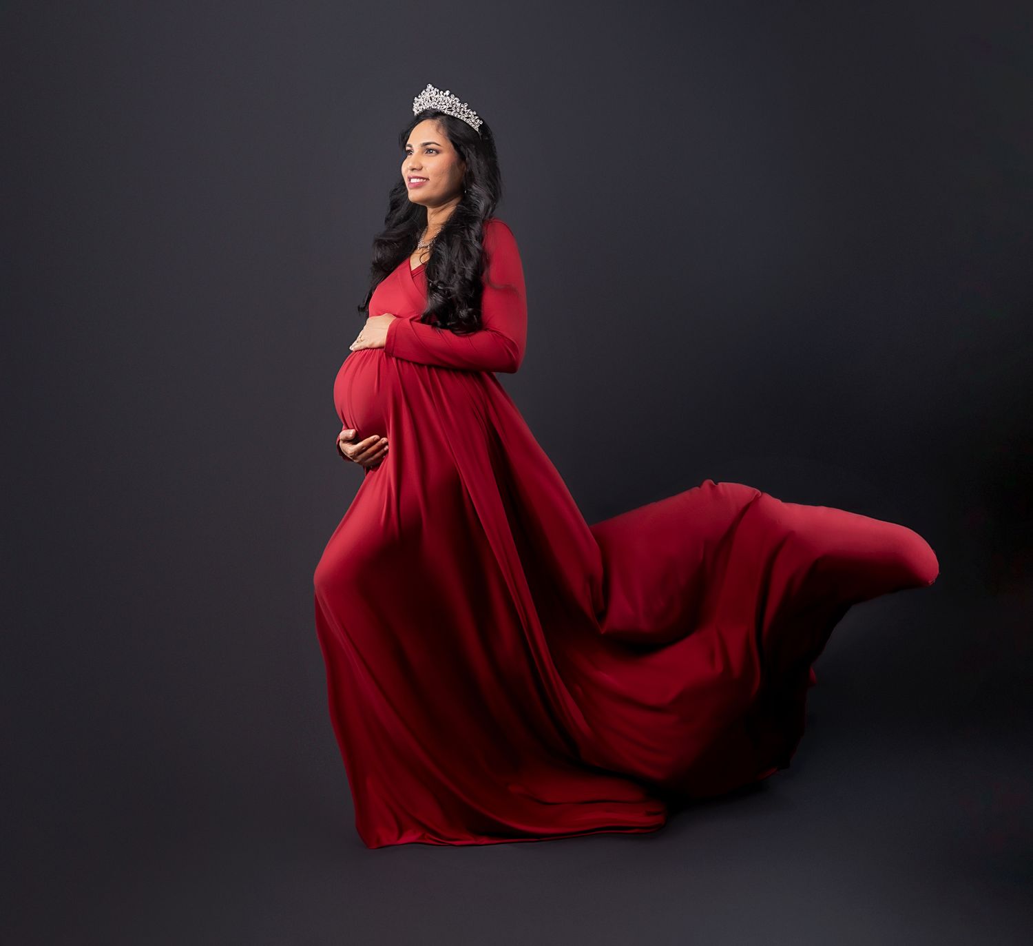 A pregnant woman wearing a flowing red maternity gown and tiara poses elegantly against a dark backdrop.