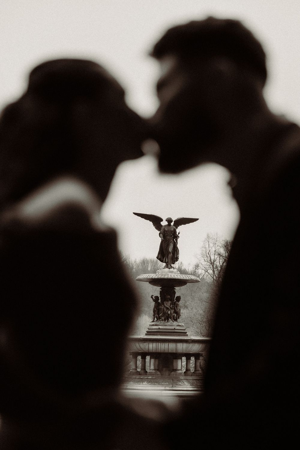 Dark silhouetted figures in the foreground frame a distant statue of an eagle monument in a soft focus black and white scene.