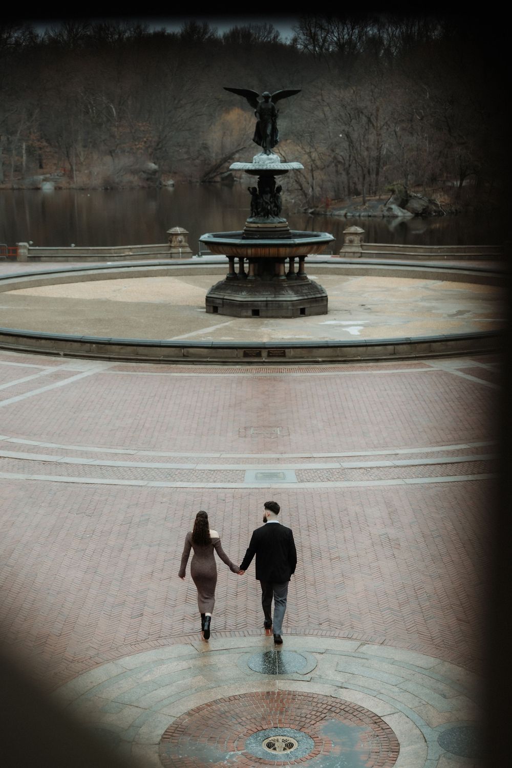 Two silhouetted figures walk away from camera across paved circle with decorative fountain in Central Park at dusk.