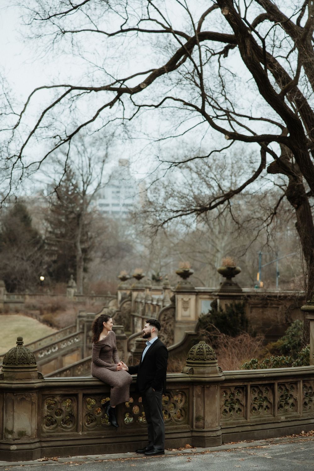 A couple sits on an ornate stone staircase in Central Park during winter with bare trees and city buildings in background.