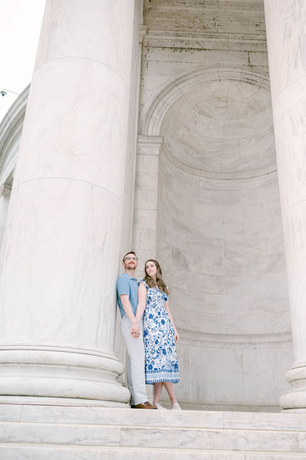 A couple poses between classic white marble columns in an elegant architectural setting.