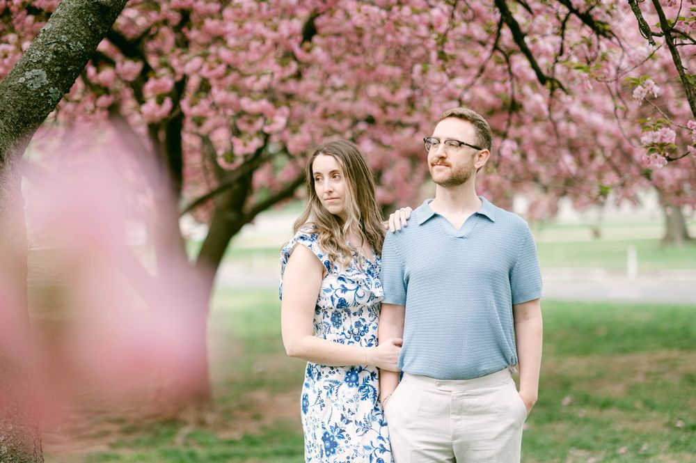 A couple stands beneath blooming pink cherry blossoms in a spring garden setting.
