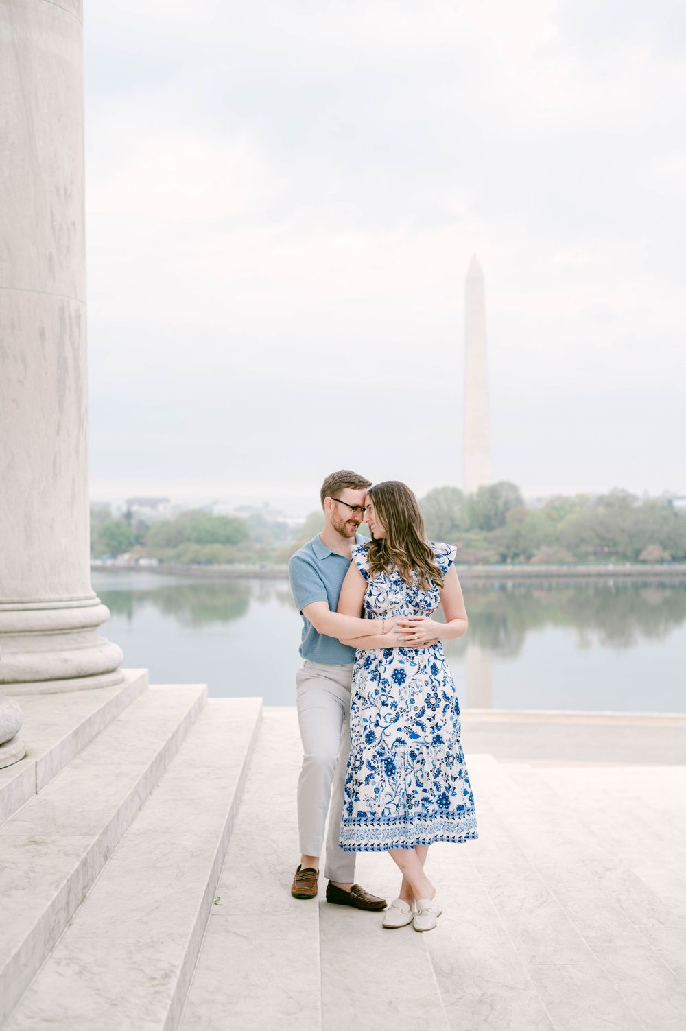 A tender embrace between a couple near marble columns with the Washington Monument and water in view.