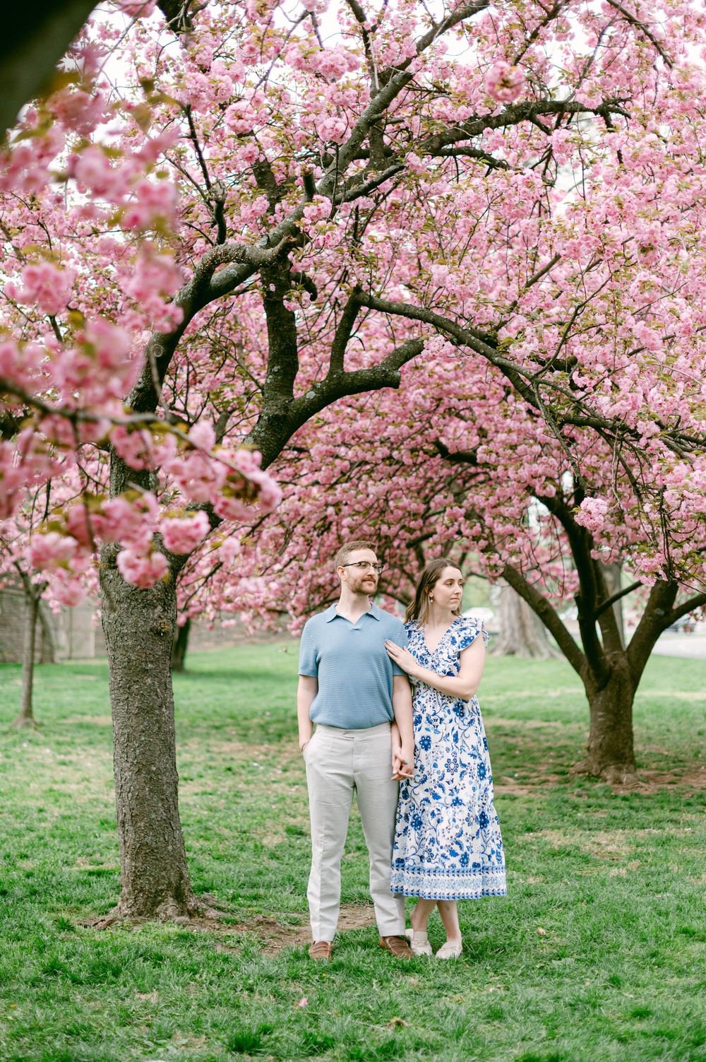 A couple stands beneath blooming pink cherry blossom trees on a spring day.