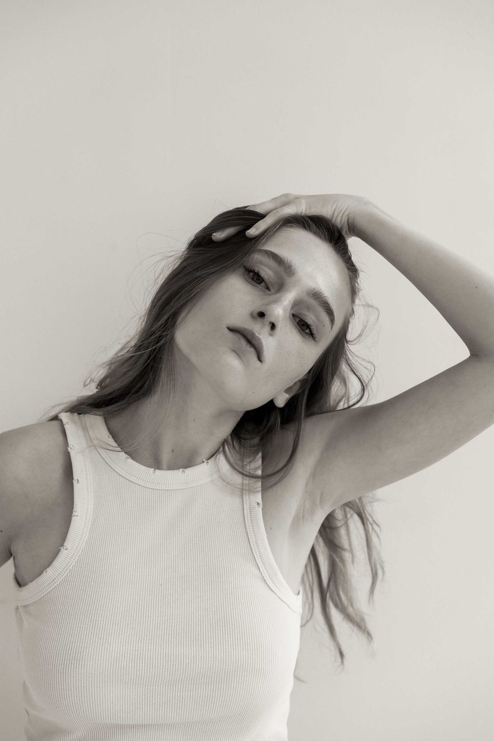 Black and white artistic portrait of someone in a white tank top striking a relaxed pose against a light background.