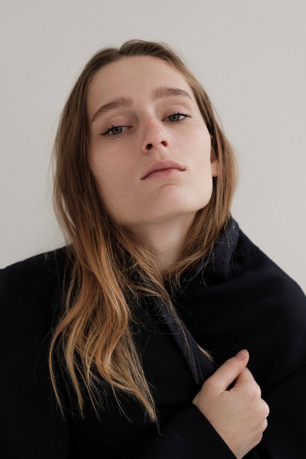 Person wearing black hoodie poses against white wall with natural light in minimalist portrait photograph.