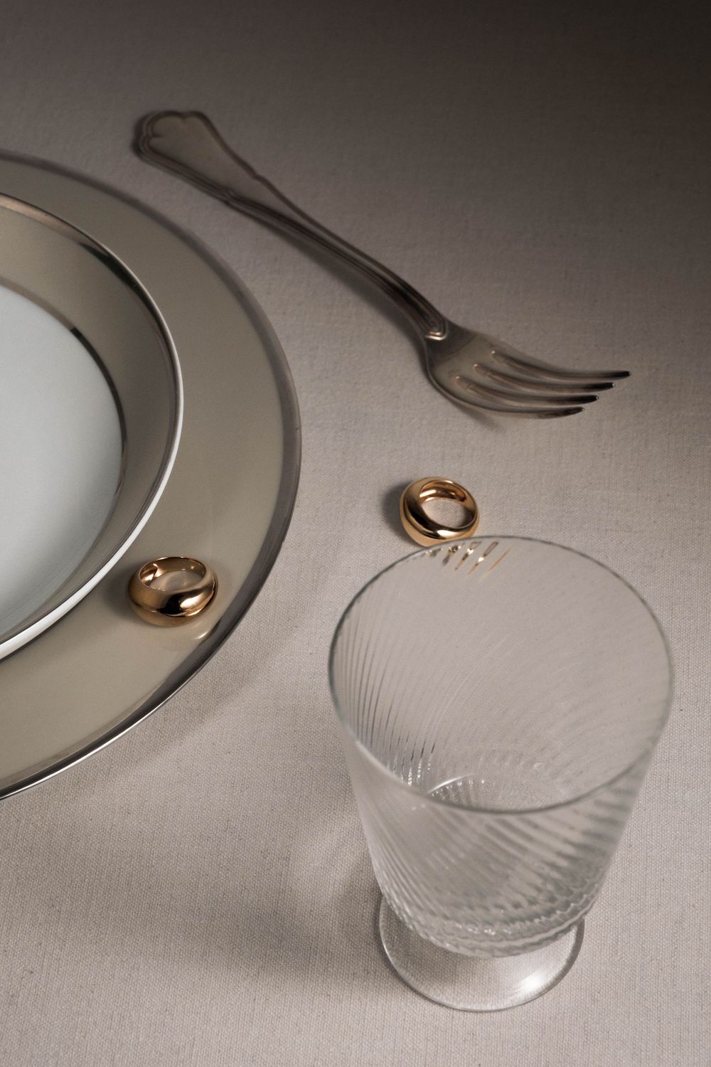Gold wedding rings sit elegantly beside white porcelain plates and crystal glassware on a neutral linen tablecloth.