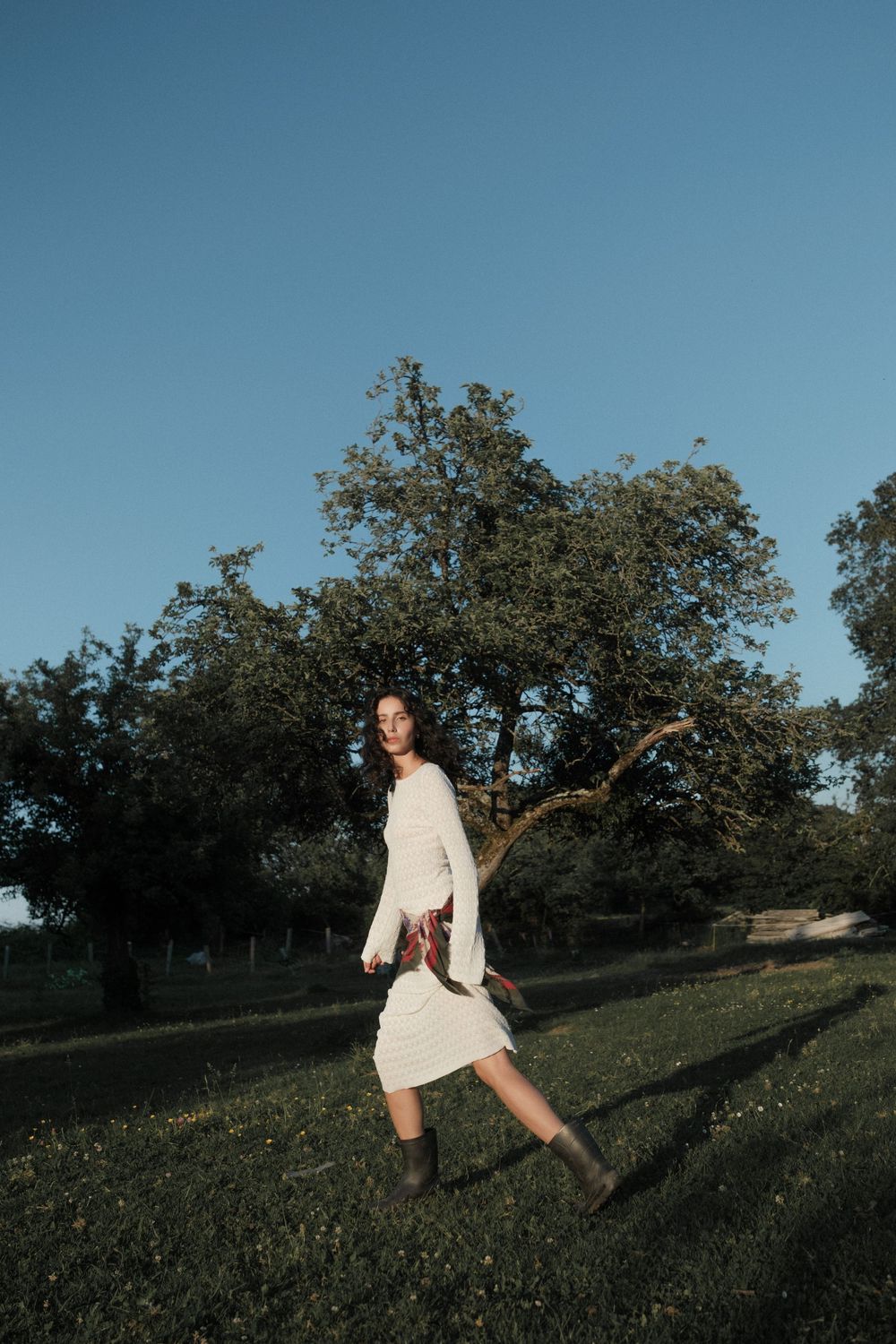 Woman in white dress and boots walking on green grass field with large tree and clear blue sky backdrop.