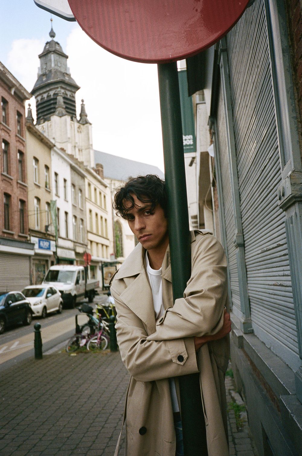 Young man in beige trench coat leaning against a green pole on a European city street with historic church tower backdrop.