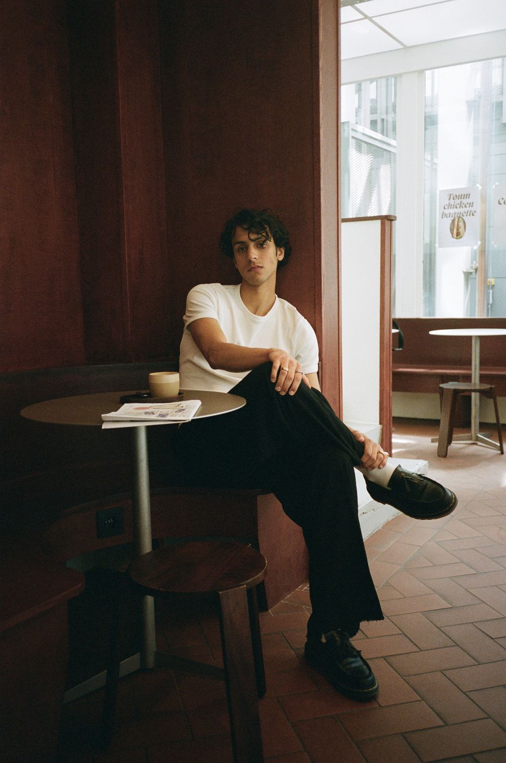 Young man in white t-shirt and black pants sitting at a small cafe table with a coffee cup.