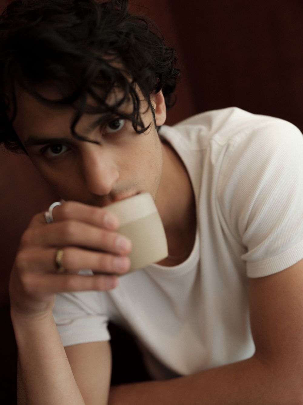 Young man with curly dark hair sipping from a small white cup, wearing a white t-shirt and silver ring.