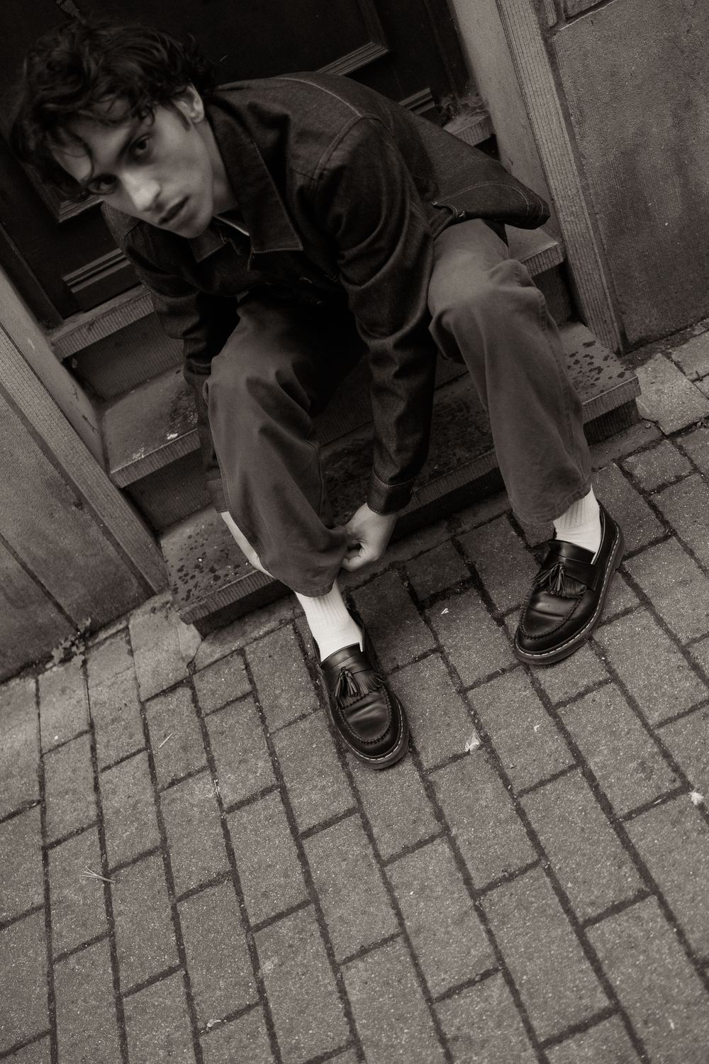 Young man in dark jacket and trousers with white socks and black loafers sitting on steps in black and white photo.