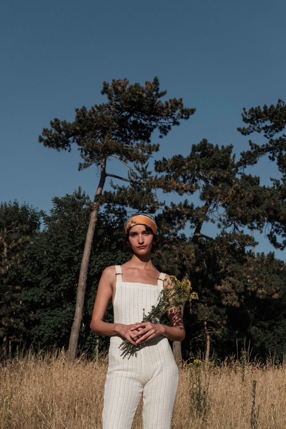 Person in white outfit poses against a backdrop of tall grass and pine trees on a sunny day.