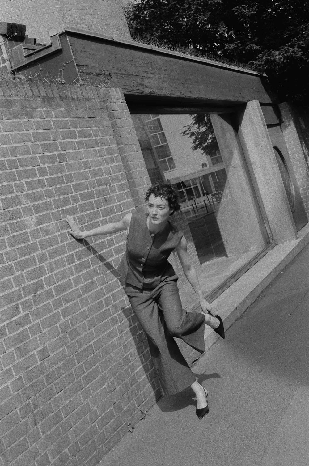 A person in casual attire leans playfully against a brick wall on a street corner in a black and white photograph.