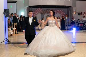 Quinceañera girl in sparkling white ball gown makes grand entrance with escort on decorated dance floor.