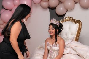 Young woman in ornate quinceañera gown and crown sits on throne chair with guest at pink balloon backdrop.