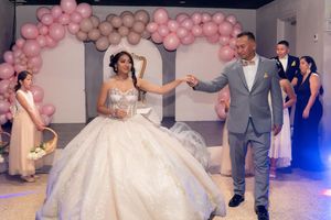 Quinceañera girl in white ball gown enters celebration hall with escort under pink balloon decorations.