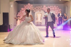 Young woman in sparkly ball gown dances with man in gray suit at quinceañera celebration with pink balloon arch.