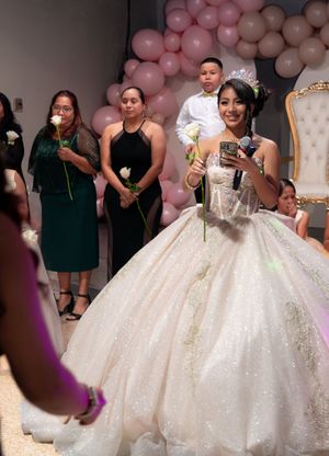 Quinceañera girl in white glittery ball gown holds microphone and rose during ceremony with guests and pink balloons.