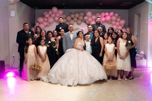 Large family group photo at quinceañera with girl in white ball gown surrounded by guests before pink balloon backdrop.