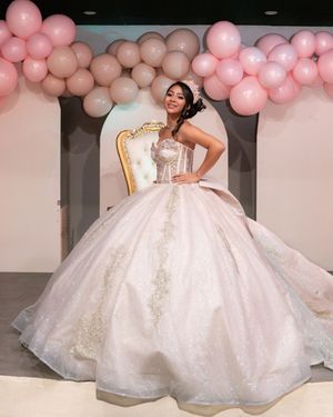 Quinceañera girl in elaborate white sparkly ball gown stands before golden throne chair with pink balloon backdrop.