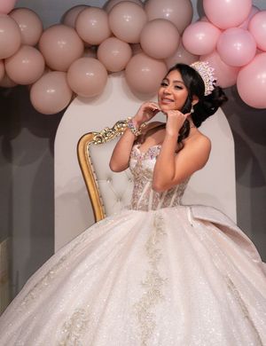 Quinceañera girl in white glittery gown and tiara sits gracefully on golden throne chair with pink balloon decor.