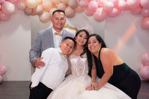 Quinceañera girl in white gown poses with family of four including father, mother, and brother before pink balloons.